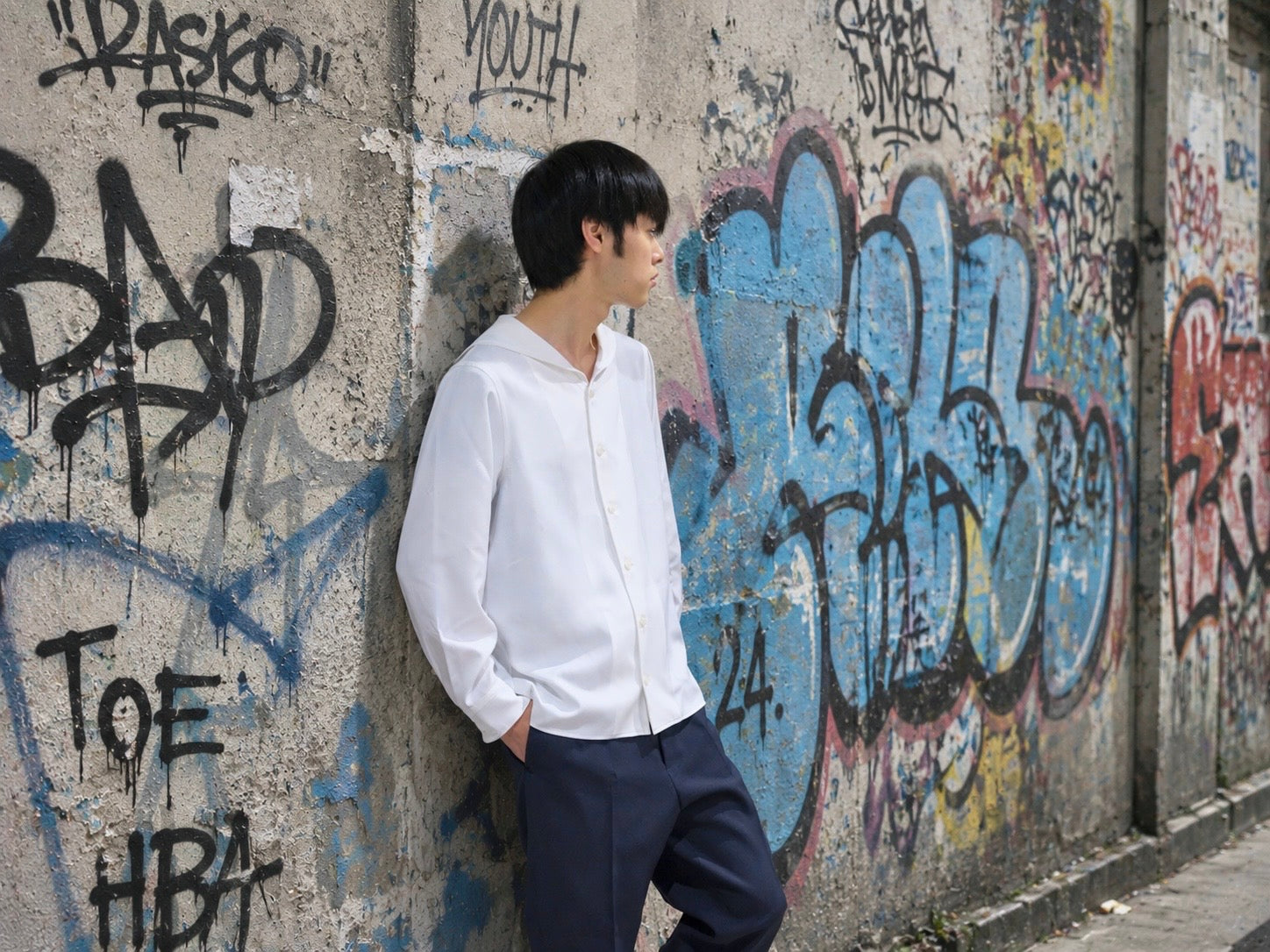白いセーラーカラーシャツとネイビーパンツを着て落書きの壁にもたれる男性モデル